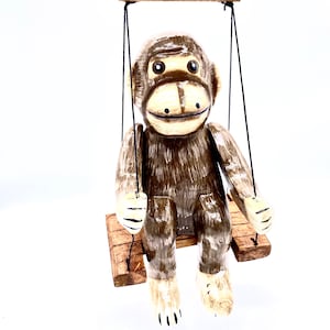Puede incluir: Una figurita de mono de madera con pelaje marrón y cara pintada está sentada en un columpio de madera. El mono tiene una amplia sonrisa y ojos grandes y redondos. El columpio está suspendido de un cordón negro.