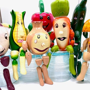 May include: A set of seven hand-painted wooden figurines of vegetables and a mushroom. Each figurine has a smiling face and is sitting on a glass jar. The vegetables include an onion, a carrot, a corn cob, a red pepper, and a green bean. The mushroom is brown with yellow accents.
