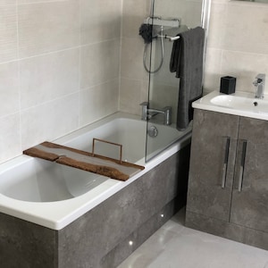 Puede incluir: Una bañera blanca con una bandeja de baño de madera, una pared de azulejos grises y un gabinete gris con un lavabo blanco y un grifo cromado.