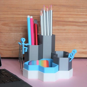 May include: A grey hexagonal desk organiser with multiple compartments for pens, pencils, and other office supplies. The organiser has a blue and white base and features small blue figures climbing the sides.