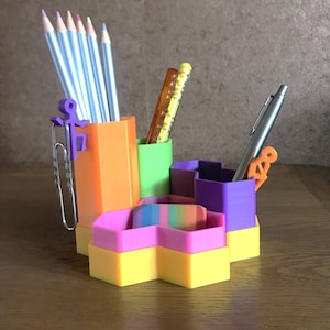 May include: A colourful hexagonal desk organiser with compartments for pens, pencils, and other office supplies. The organiser is made of plastic and has a bright yellow, pink, green, purple, and orange colour scheme.