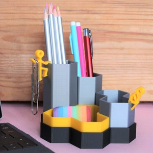 May include: A grey hexagonal desk organiser with multiple compartments for pens, pencils, and other office supplies. The organiser has a yellow honeycomb design and features a small yellow figure on the side.