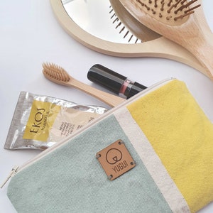 May include: A light blue and yellow canvas zippered pouch with a brown leather tag that says "YUGUI". The pouch is open and contains a small package of face masks, a wooden toothbrush, and a lipstick.