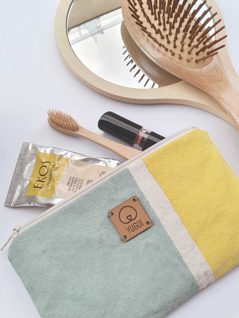 May include: A light blue and yellow canvas zippered pouch with a brown leather tag that says "YUGUI". The pouch is open and contains a small package of face masks, a wooden toothbrush, and a lipstick.