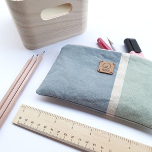 May include: A blue and green striped canvas pencil case with a brown leather patch that says "YUGUI". The case is open and has pencils, a ruler, and a pink highlighter inside.