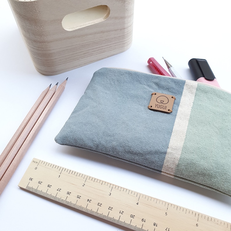 May include: A blue and green striped canvas pencil case with a brown leather patch that says "YUGUI". The case is open and has pencils, a ruler, and a pink highlighter inside.