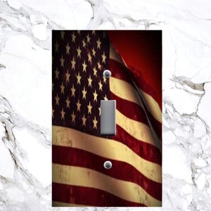 May include: A light switch cover featuring a faded American flag with red, white, and blue stripes and stars. The flag is draped over a red background.