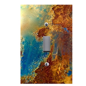 May include: A decorative light switch cover with an abstract design. The cover features a blend of rusty brown, blue, and yellow hues, with a metallic silver switch and two silver screws. The design has a textured, weathered appearance.