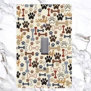 May include: Light switch cover with a beige background and a pattern of brown, black, and gray paw prints, bones, and the word "woof" repeated throughout.