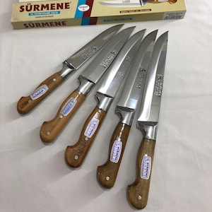 Puede incluir: Seis cuchillos de carnicero plateados con mangos de madera. Los cuchillos están dispuestos en forma de abanico. Los cuchillos tienen el nombre de la marca "SÜRMENE" impreso en las hojas y los mangos. Los cuchillos están empaquetados en una caja con el nombre de la marca "SÜRMENE" impreso en la caja.