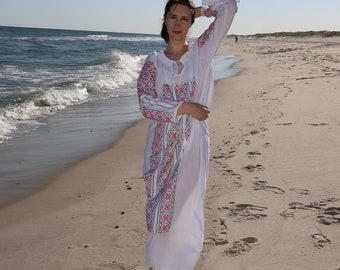 May include: A white embroidered dress with long sleeves and a flowing skirt. The sleeves and front panel feature a detailed floral pattern in red, pink, and black. The dress is worn on a sandy beach with the ocean in the background.