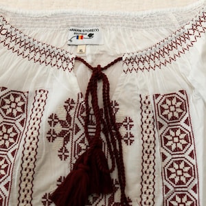 May include: White blouse with burgundy embroidery. The blouse features a detailed floral and geometric pattern around the neckline and on the front. It has a tassel tie and the tag reads "HANIAN STORE(Y) BOUTIQUE".