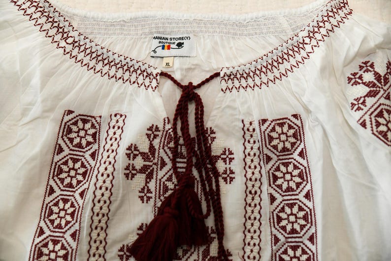 May include: White blouse with burgundy embroidery. The blouse features a detailed floral and geometric pattern around the neckline and on the front. It has a tassel tie and the tag reads "HANIAN STORE(Y) BOUTIQUE".