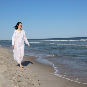 May include: A woman in a white, long-sleeved dress walks along a sandy beach. The dress has a vertical striped pattern. The sky is clear blue, and the ocean waves are visible in the background.