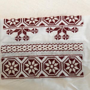 May include: Close-up of white fabric with intricate maroon embroidery. The design features geometric and floral patterns, including repeating star-like shapes and decorative borders. The embroidery is detailed and appears to be hand-stitched, showcasing traditional craftsmanship.