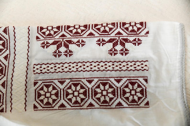 May include: Close-up of white fabric with intricate maroon embroidery. The design features geometric and floral patterns, including repeating star-like shapes and decorative borders. The embroidery is detailed and appears to be hand-stitched, showcasing traditional craftsmanship.
