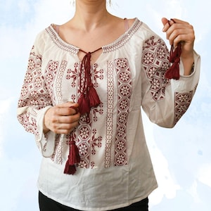 May include: A white blouse with long sleeves and detailed burgundy embroidery. The blouse has a V-neck with a tassel tie and decorative patterns on the sleeves and bodice. The sleeves are long and loose.