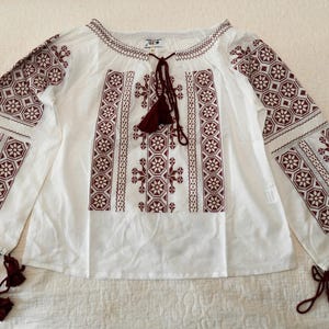 May include: White blouse with burgundy embroidery. The long-sleeved top features intricate floral and geometric patterns on the sleeves and bodice. It has a round neckline with a tassel tie and tassels at the end of the sleeves. The blouse is laid flat on a white surface.