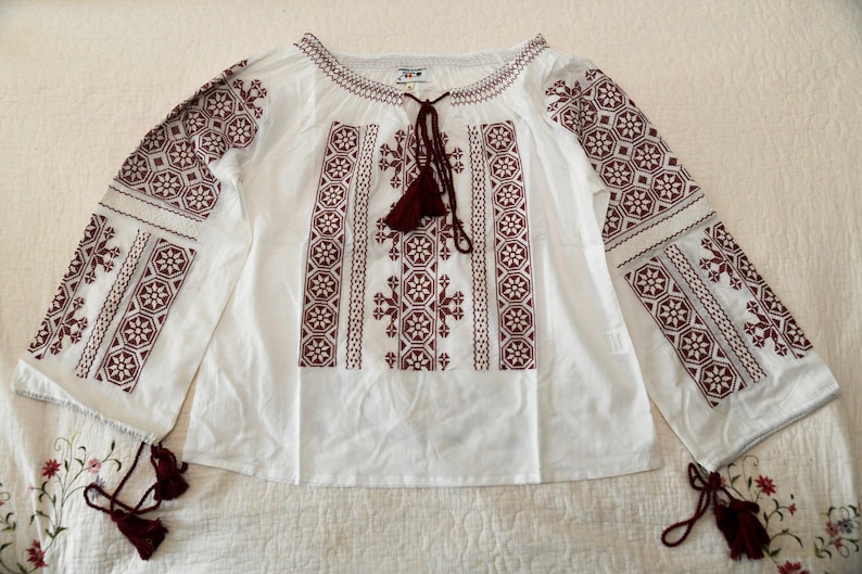 May include: White blouse with burgundy embroidery. The long-sleeved top features intricate floral and geometric patterns on the sleeves and bodice. It has a round neckline with a tassel tie and tassels at the end of the sleeves. The blouse is laid flat on a white surface.
