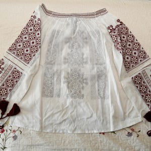 May include: White off-the-shoulder blouse with long sleeves. The sleeves and neckline are adorned with burgundy geometric embroidery. The blouse features a loose, flowing design and decorative tassels at the cuffs. A traditional, ethnic style.