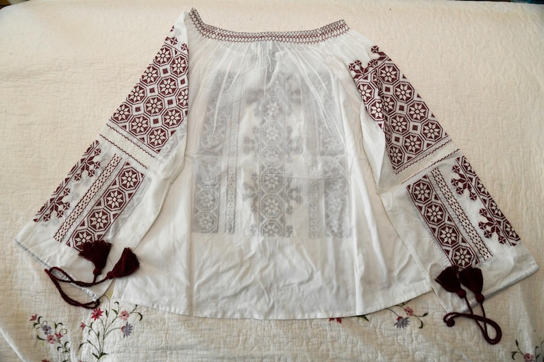 May include: White off-the-shoulder blouse with long sleeves. The sleeves and neckline are adorned with burgundy geometric embroidery. The blouse features a loose, flowing design and decorative tassels at the cuffs. A traditional, ethnic style.