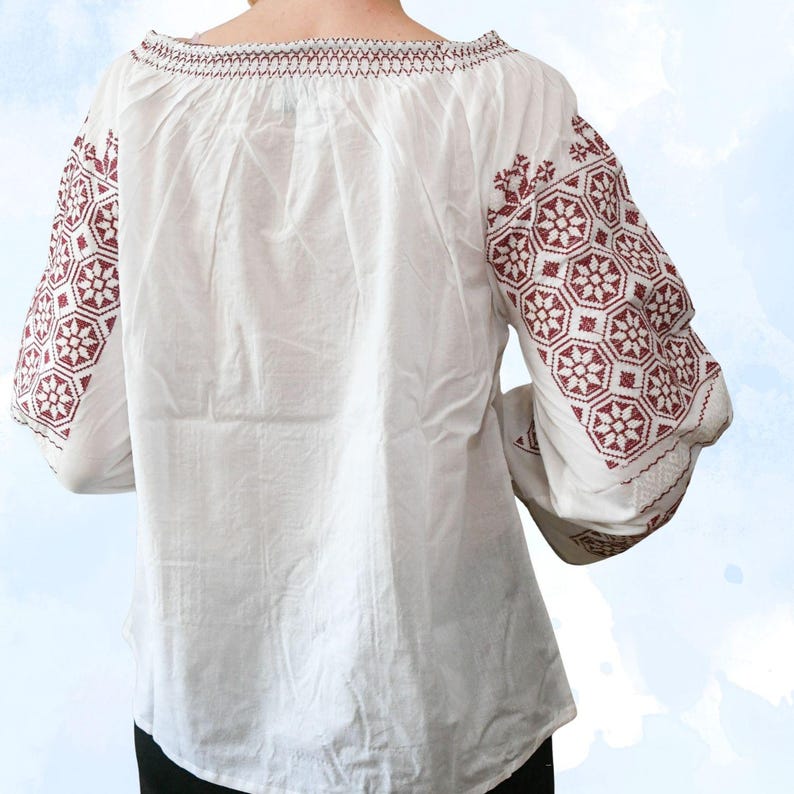May include: White blouse with burgundy floral embroidery on the sleeves and neckline. The long sleeves have a slight bell shape. The embroidery features geometric and floral patterns. The blouse is made of a lightweight fabric.