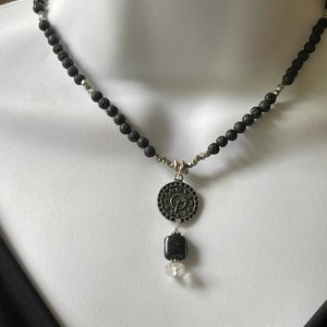 May include: A black lava rock necklace with a silver clock pendant and a black bead. The clock face has Roman numerals and a silver hand.