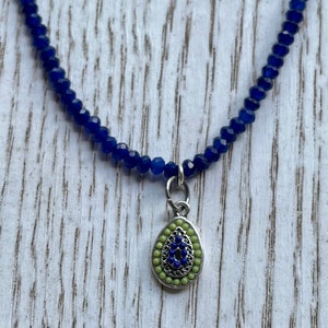 May include: A blue beaded necklace with a silver charm featuring a blue evil eye with green accents.