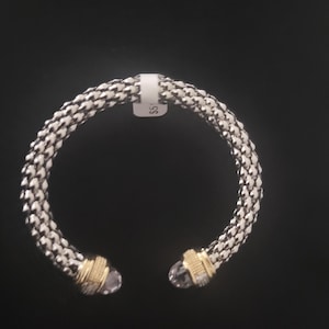 Puede incluir: Una pulsera tejida plateada y blanca con detalles dorados y extremos de cristal transparente. La pulsera está abierta en los extremos y tiene una etiqueta. La pulsera está sobre un fondo negro.