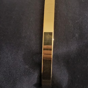 May include: A gold-coloured, rectangular bangle bracelet. The bracelet has a smooth, polished surface and a simple, elegant design. The bracelet is shown on a dark blue fabric background.
