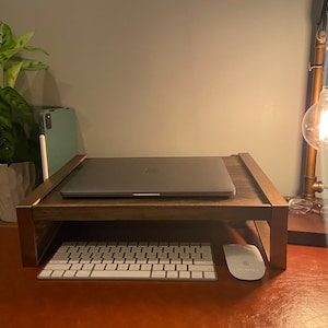 May include: A dark wood laptop stand with copper-colored metal accents. A closed laptop sits on the stand, with a white keyboard and mouse below. A desk lamp and a small potted plant are also visible.