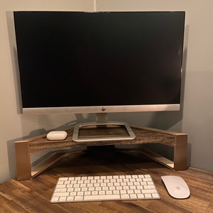 May include: A gold metal monitor stand with a wooden top, a white wireless keyboard, and a white wireless mouse on a wooden desk.