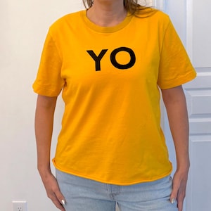 Puede incluir: Una camiseta de color amarillo mostaza con la palabra "YO" impresa en negro. La camiseta tiene mangas cortas y cuello redondo. La persona lleva vaqueros azul claro.