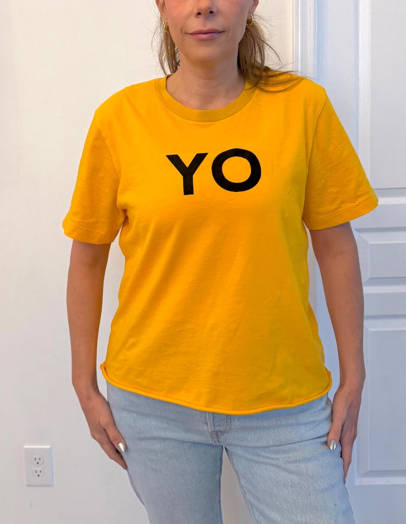 Puede incluir: Una camiseta de color amarillo mostaza con la palabra "YO" impresa en negro. La camiseta tiene mangas cortas y cuello redondo. La persona lleva vaqueros azul claro.