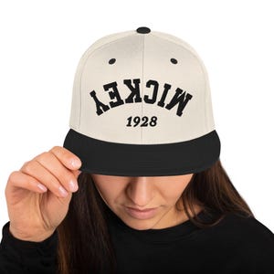 Könnte beinhalten: Eine beige und schwarze Baseballkappe mit dem Wort "MICKEY" in schwarzen Buchstaben auf der Vorderseite. Das Jahr "1928" ist unter dem Wort gedruckt.