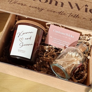May include: A gift box with a candle and a glass match bottle. The candle label reads "Keanan and Shauna" and the date "15.04.2025". A card reads "Custom Wicks". The box is filled with shredded paper.