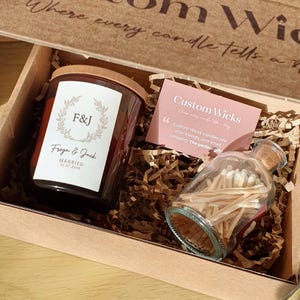 May include: A gift box with a candle and a match bottle. The candle has a label with the initials "F&J", the names "Freya & Jack", and the wedding date. The box also includes a card that says "Custom Wicks".