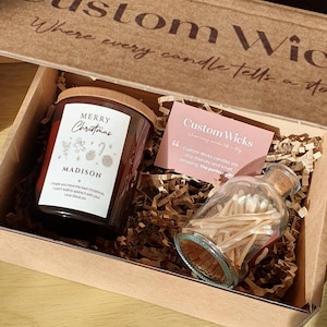 May include: A brown cardboard box with its lid open, revealing a Christmas-themed candle with a brown lid and a glass jar filled with matches. The candle label reads "Merry Christmas" and "Madison". The box also includes a card that reads "Custom Wicks" and "Custom Wicks candles are eco-friendly and smell amazing. The perfect gift!"