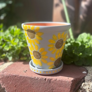 May include: A gray terracotta planter with a painted design of yellow sunflowers. The planter has a matching saucer with a dotted pattern.
