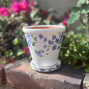 May include: White ceramic flower pot with a blue and green floral design. The pot has a saucer.
