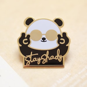 May include: Gold-tone enamel pin featuring a black and white panda wearing sunglasses and making a peace sign with each hand. The text "Stay Shady" is written below the panda.