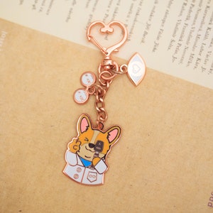 May include: Rose gold keychain featuring a cartoon dog wearing a white lab coat and holding a microphone. The keychain also includes a heart charm and a charm with an eye and a heart.