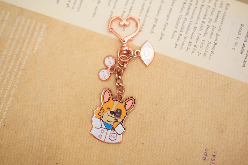 May include: Rose gold keychain featuring a cartoon dog wearing a white lab coat and holding a microphone. The keychain also includes a heart charm and a charm with an eye and a heart.