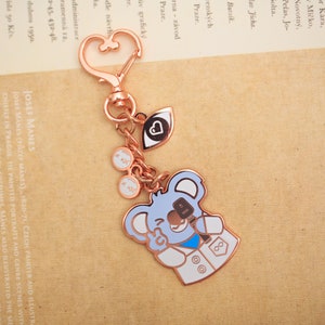 May include: A rose gold keychain with a koala bear charm. The koala is wearing a white lab coat with a blue stethoscope and a blue eye patch. The keychain also has a heart charm, an eye charm, and a pair of glasses charm.