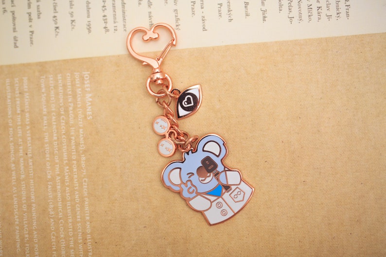 May include: A rose gold keychain with a koala bear charm. The koala is wearing a white lab coat with a blue stethoscope and a blue eye patch. The keychain also has a heart charm, an eye charm, and a pair of glasses charm.