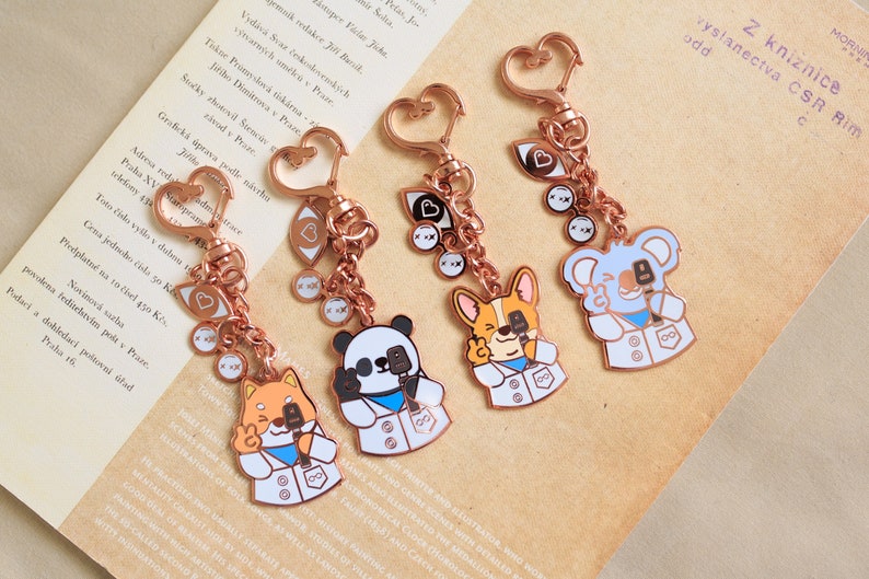 May include: Four rose gold keychains with different animal charms. Each charm is wearing a white lab coat and holding a microphone. The animals are a panda, a koala, a corgi, and a fox. The keychains also have a heart-shaped eye charm and a small round charm.