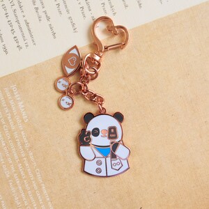 May include: A rose gold panda keychain with a microphone and a white lab coat. The keychain also features a heart, two eyes, and two small charms with the letter 'x' on them.