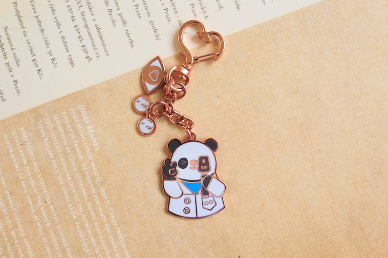 May include: A rose gold panda keychain with a microphone and a white lab coat. The keychain also features a heart, two eyes, and two small charms with the letter 'x' on them.