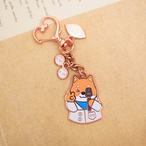 May include: A rose gold keychain featuring a cartoon Shiba Inu wearing a white lab coat and holding a microphone. The keychain also includes a heart-shaped charm and two small charms with the text "XX" on them.