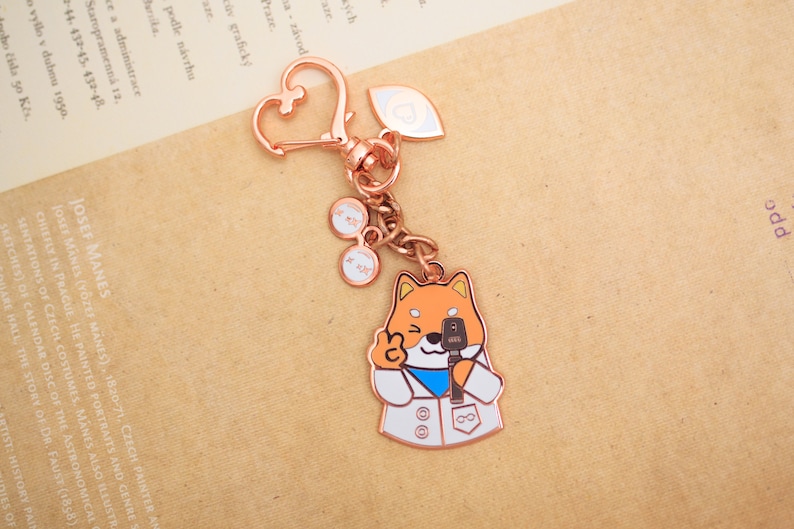 May include: A rose gold keychain featuring a cartoon Shiba Inu wearing a white lab coat and holding a microphone. The keychain also includes a heart-shaped charm and two small charms with the text "XX" on them.